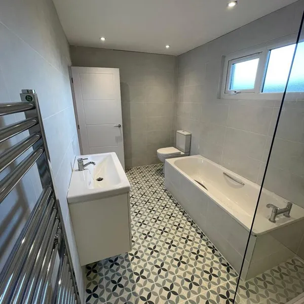 The same bathroom from the door, with a wall-hung vanity, a chrome heated towel rail and the bath under the window