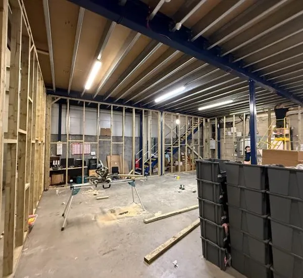 A long run of new stud framing along one side of the warehouse, with a mitre saw set up on the floor and stacked crates beside it