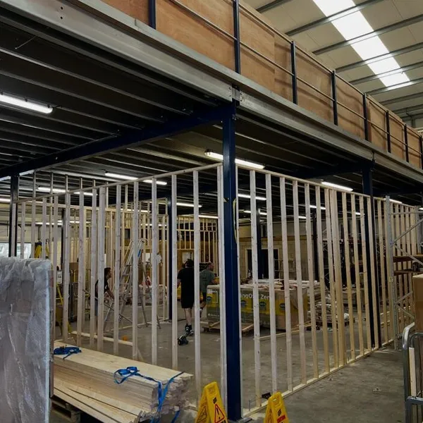 Timber stud walls going up beneath the steel mezzanine, with a pack of new timber on a pallet and wet floor signs in front