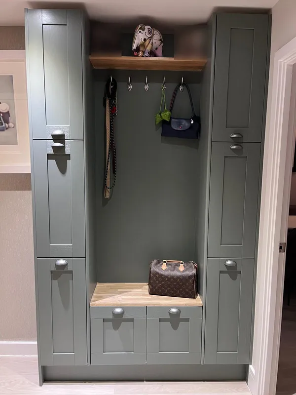 Custom-built hallway unit in sage green, with tall cupboards either side of an open bay, coat hooks under an oak shelf, and an oak bench over two drawers